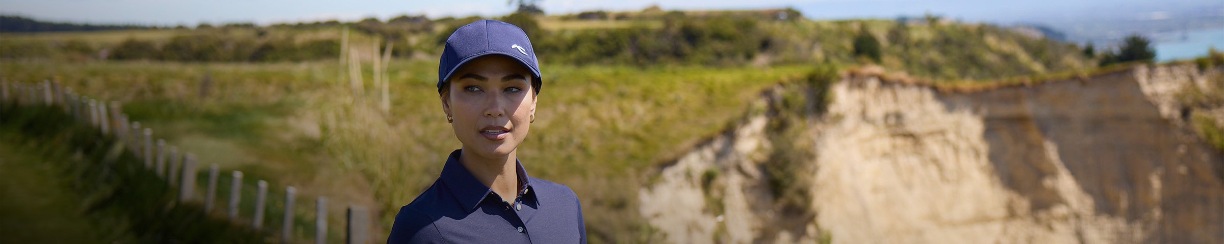 A woman wearing a KJUS cap looks out over the golf course, large cliffs and vegetation behind her.